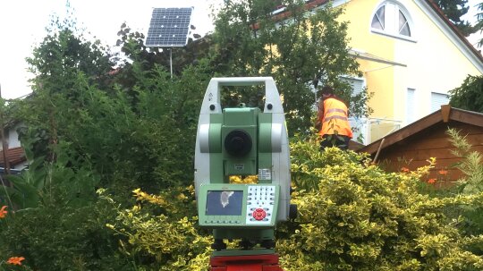 Tachymeter vor einem großen Busch stehend im Hintergrund ein Mann auf einem Garagendach stehend